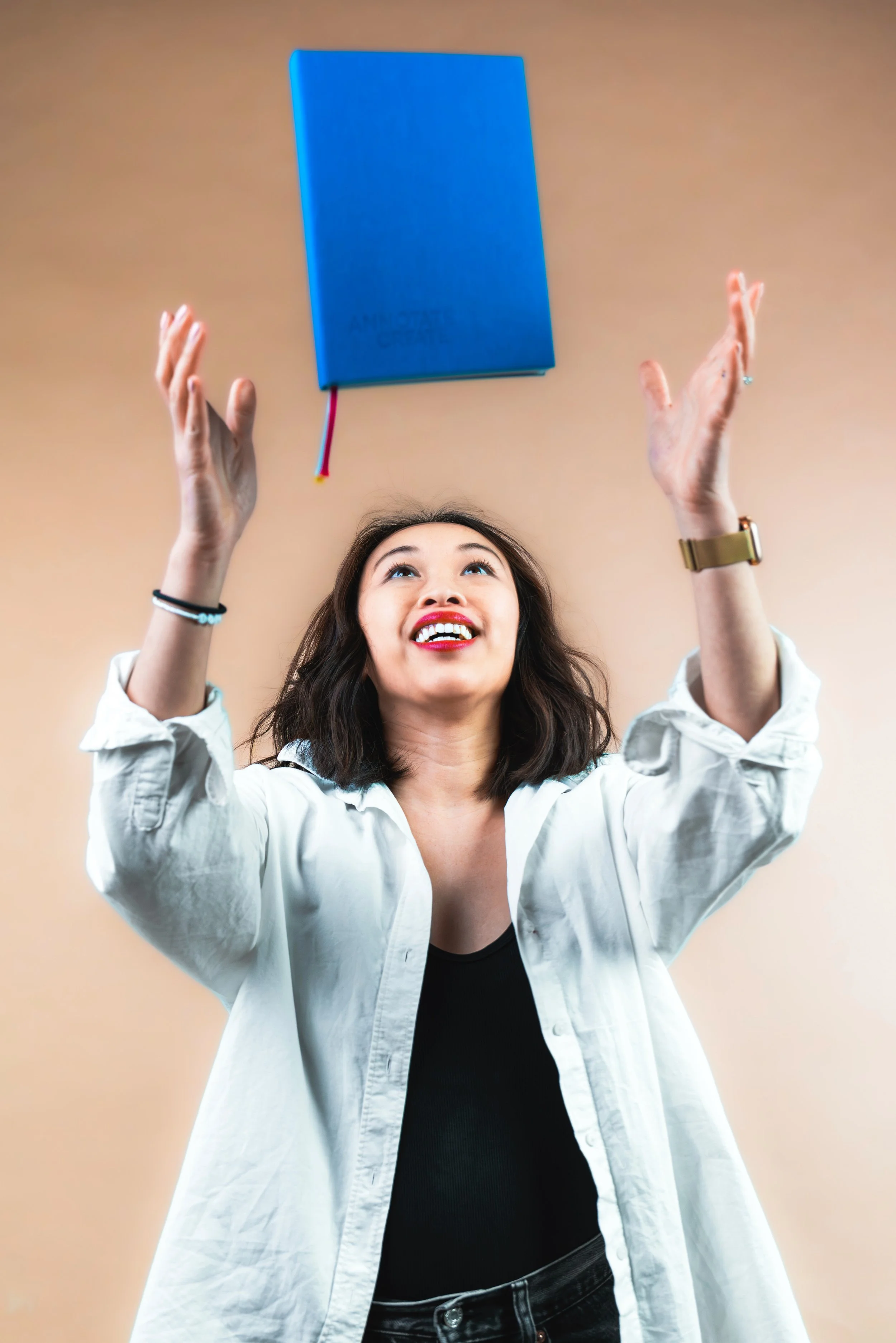 A woman with dark hair and red lipstick is smiling and raising her hands toward a blue notebook floating above her head. The background is beige.