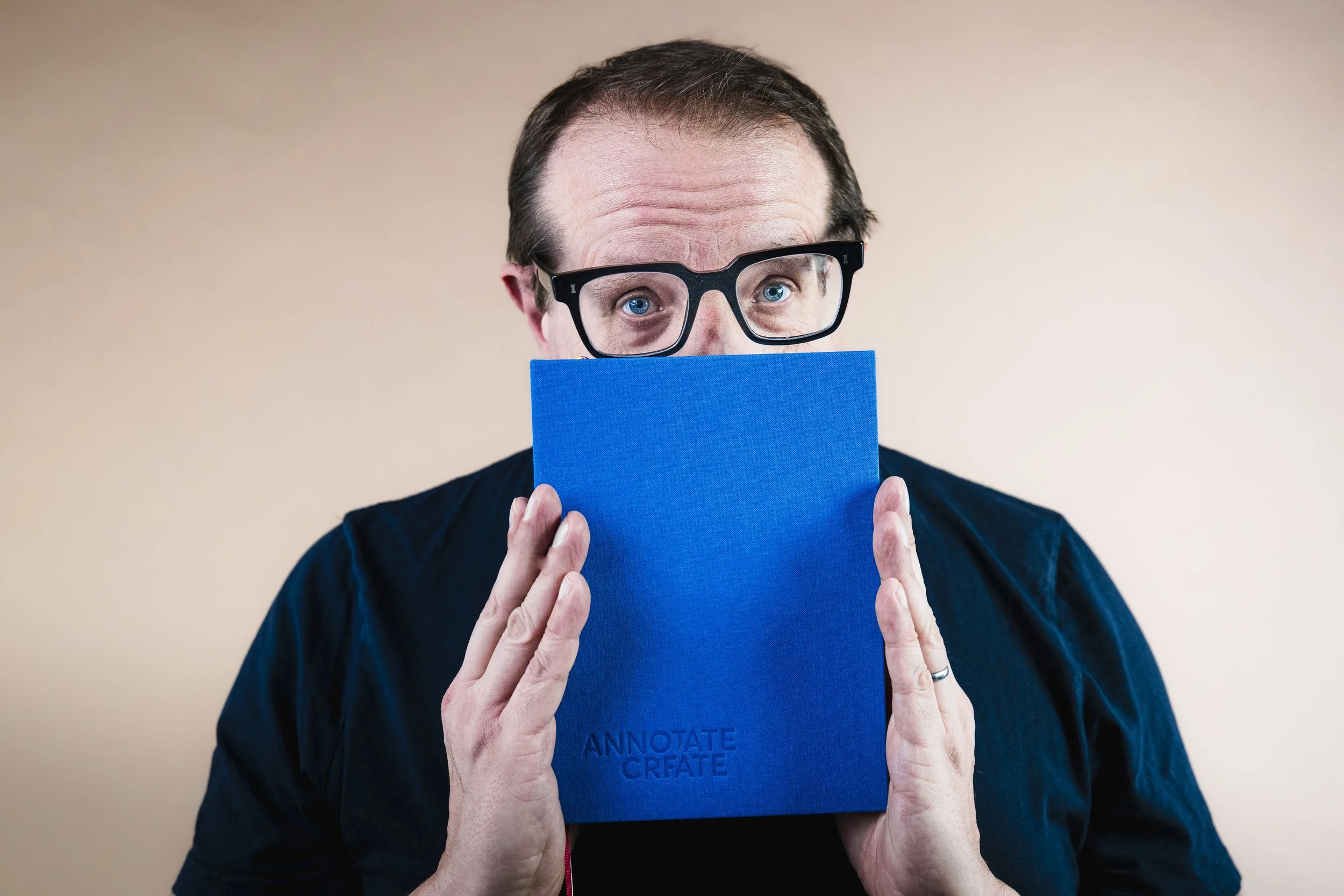 A man with glasses holding a blue notebook in front of his face, with only his eyes and hair visible.