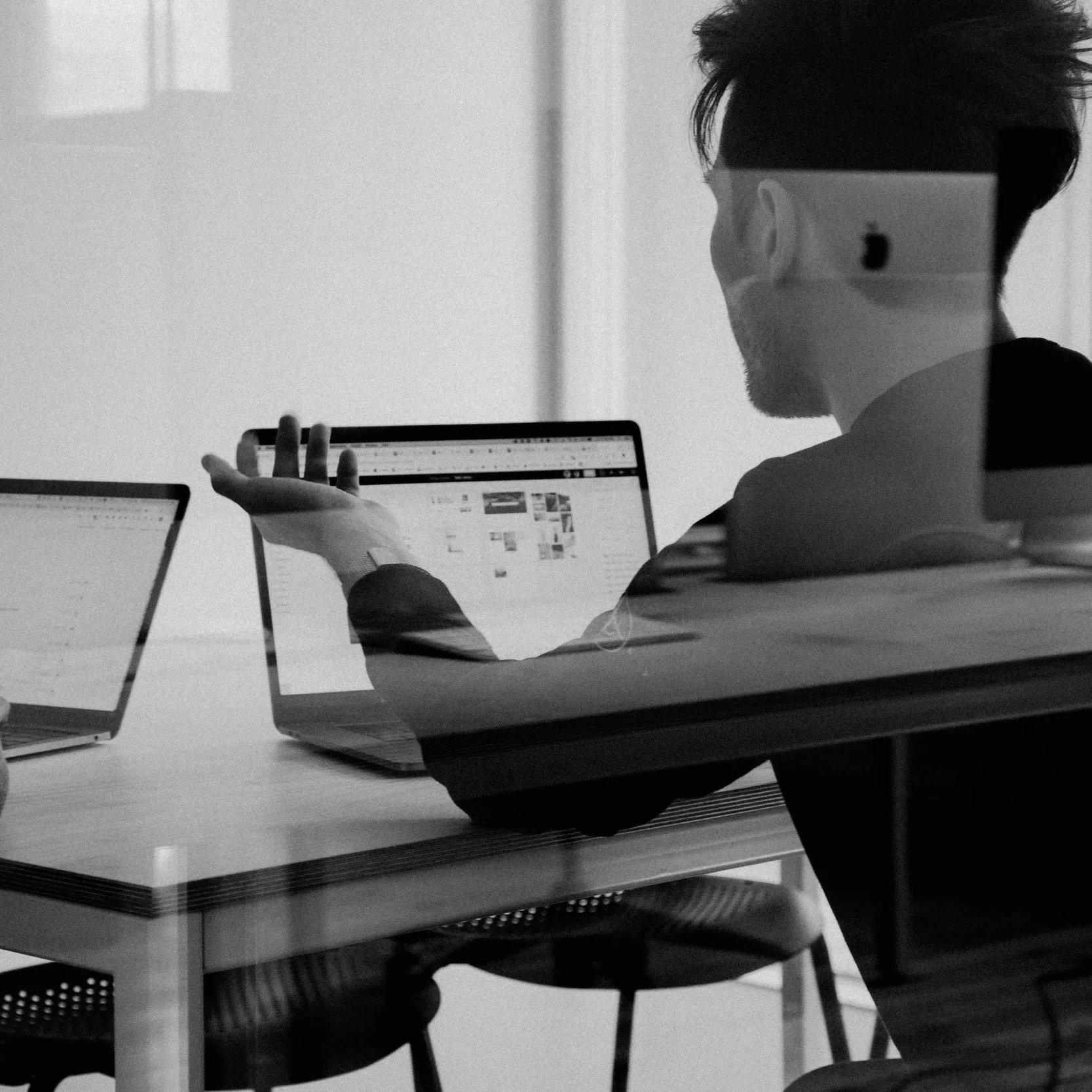 A man sitting at a desk, viewed through a glass reflection, working on a laptop with multiple windows open, in a modern office setting.