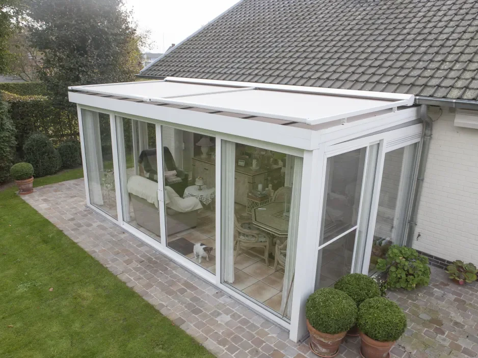 A white enclosed patio or sunroom with large sliding glass doors and windows, attached to a house with a sloped roof, surrounded by a brick patio and garden with potted plants and greenery.