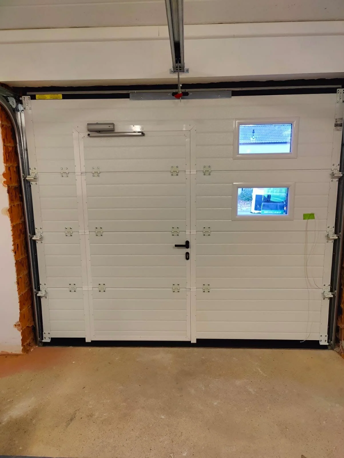 A white sectional garage door with two small windows, a black handle, and an automatic opener above. The door is in an unfinished garage with brick wall on the left and a concrete floor.