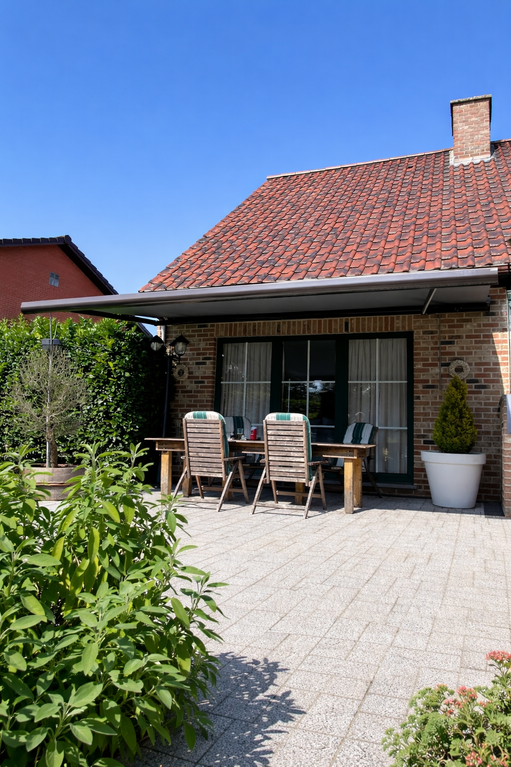 Backyard patio with wooden table and chairs, large potted plant, garden plants, brick house with a tiled roof, and a blue sky.