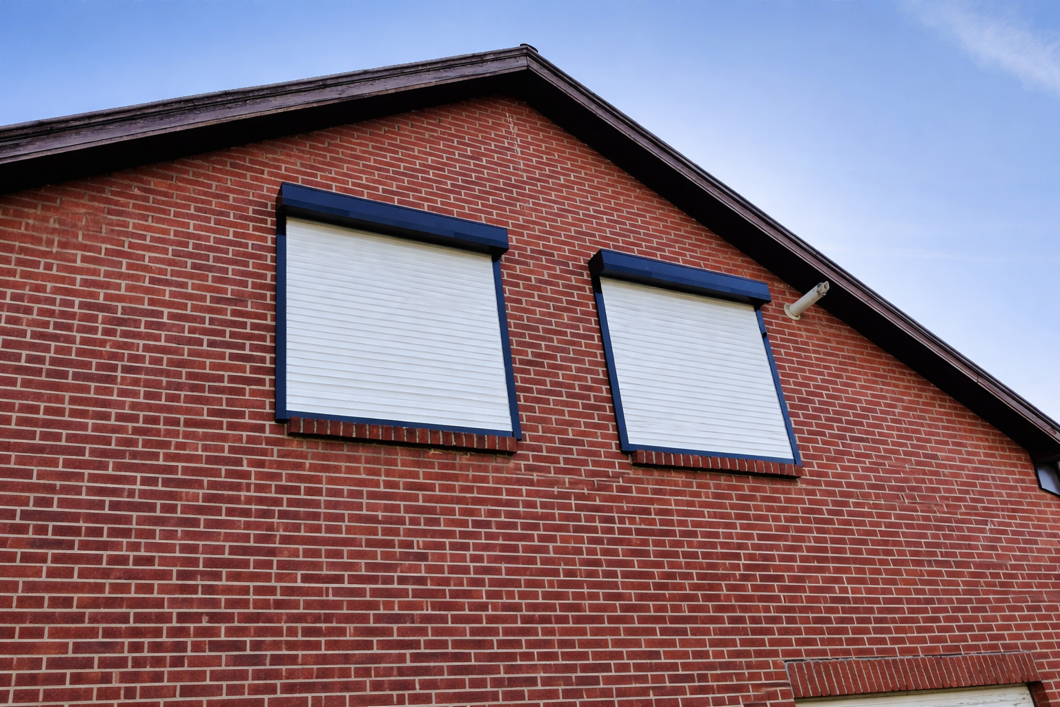 Close-up view of a brick house with two windows covered by white shutters, set against a blue sky.