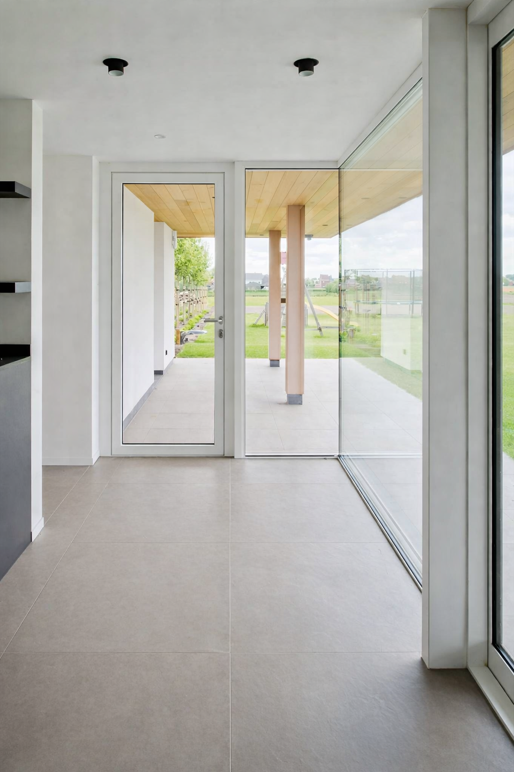 View of a modern interior space with beige tiled floors, white walls, and large glass sliding doors leading to a patio with a grassy backyard and playground.