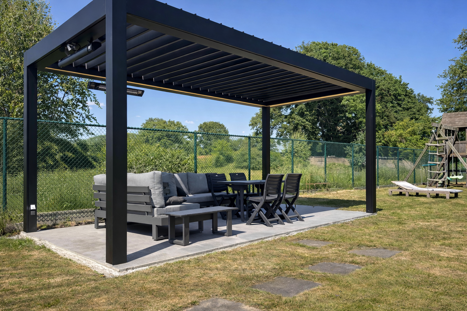 Outdoor patio area with a modern black pergola, gray outdoor furniture, a dining table with chairs, and a children's wooden playset in the background, surrounded by a chain-link fence and lush greenery under a blue sky.