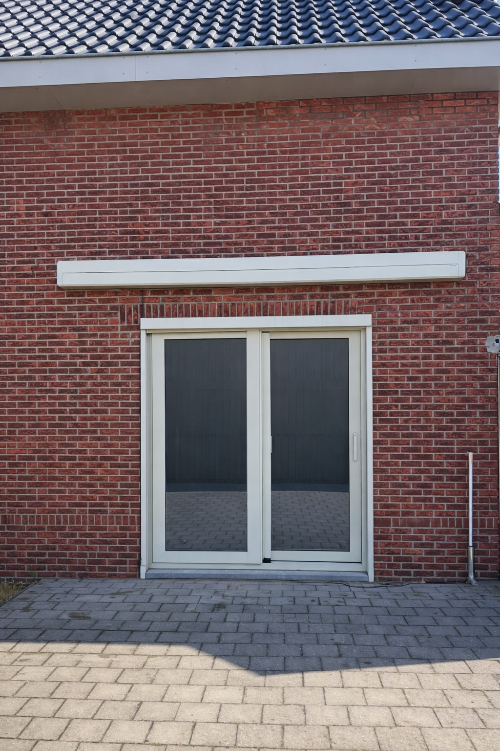 A brick building exterior features a sliding glass door, a horizontal white vent or panel above the door, and an object resembling a pipe or rod to the right of the door. The ground is paved with gray bricks.