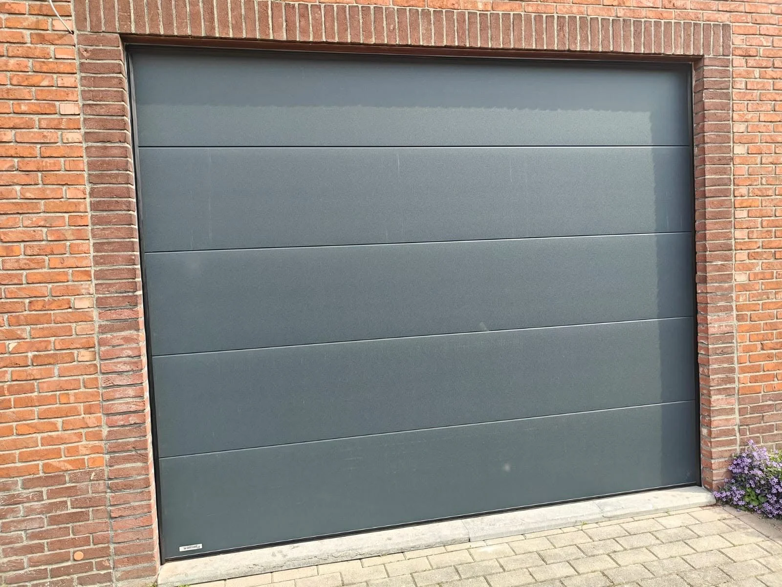 Gray garage door framed by red brick wall, with purple flowers on the right side near the bottom.