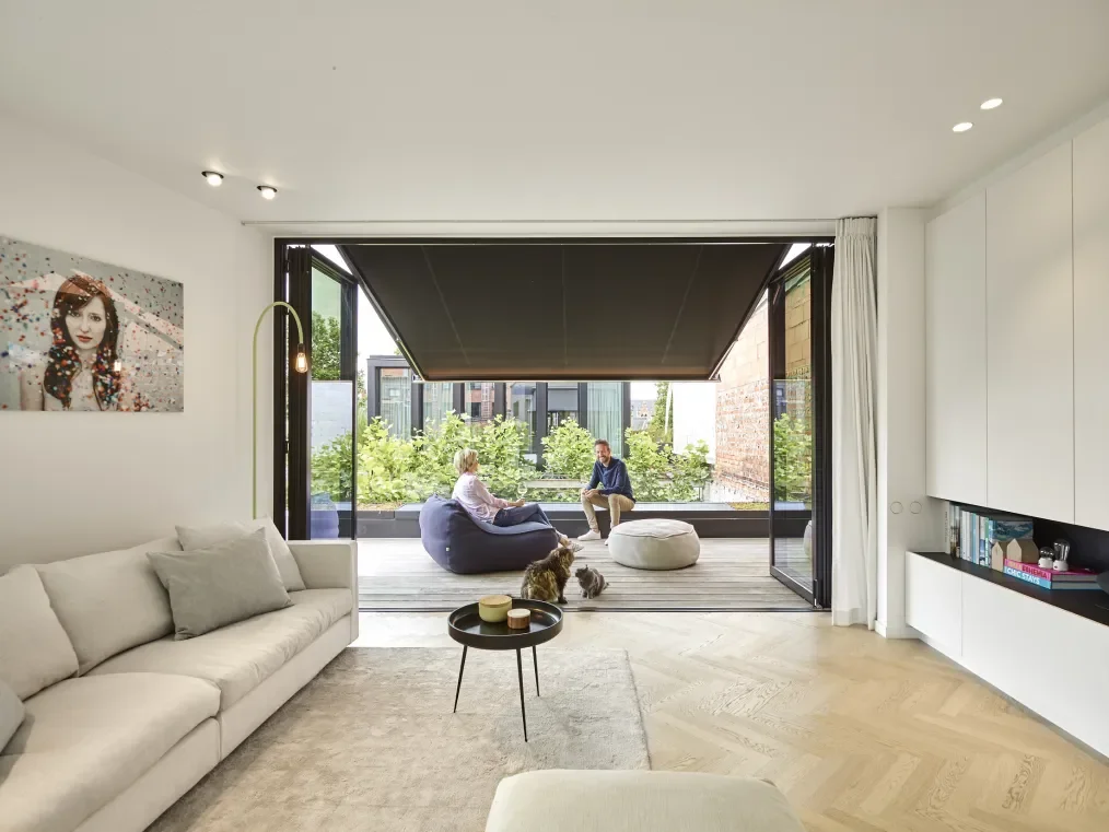 Modern living room with open sliding glass doors leading to a balcony with two people sitting and talking, and a dog sitting on the floor.