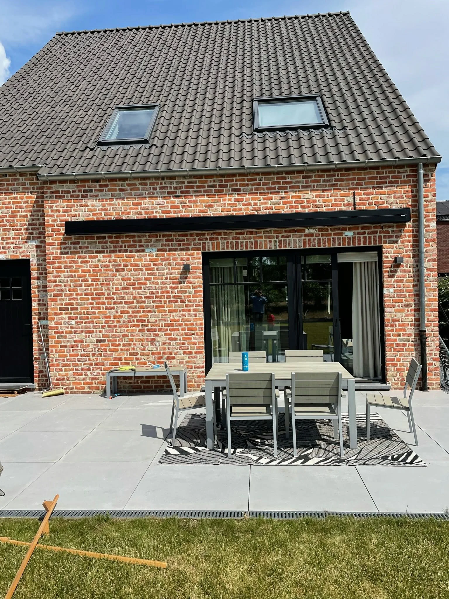 Backyard patio area with a table and chairs, brick house with black-framed sliding glass door, tiled roof with two skylights, and a grassy lawn in the foreground.