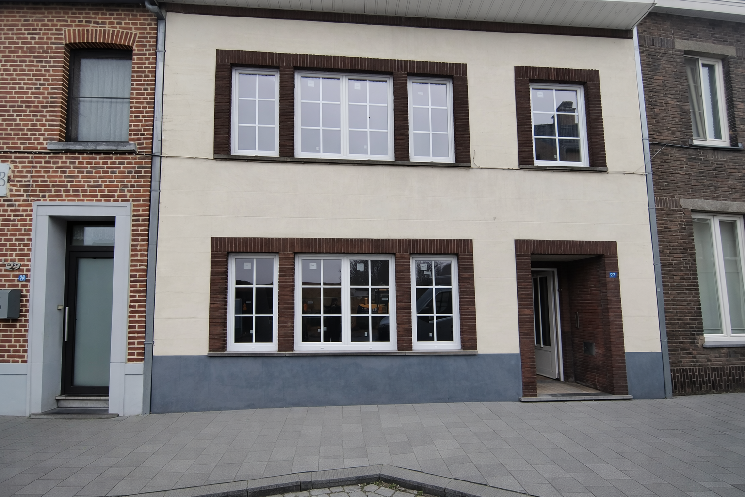 Front view of a two-story residential building with a beige facade, large white-framed windows, brick accents around the windows and door, and a small entryway with a door to the right. There is a sidewalk in front of the building.