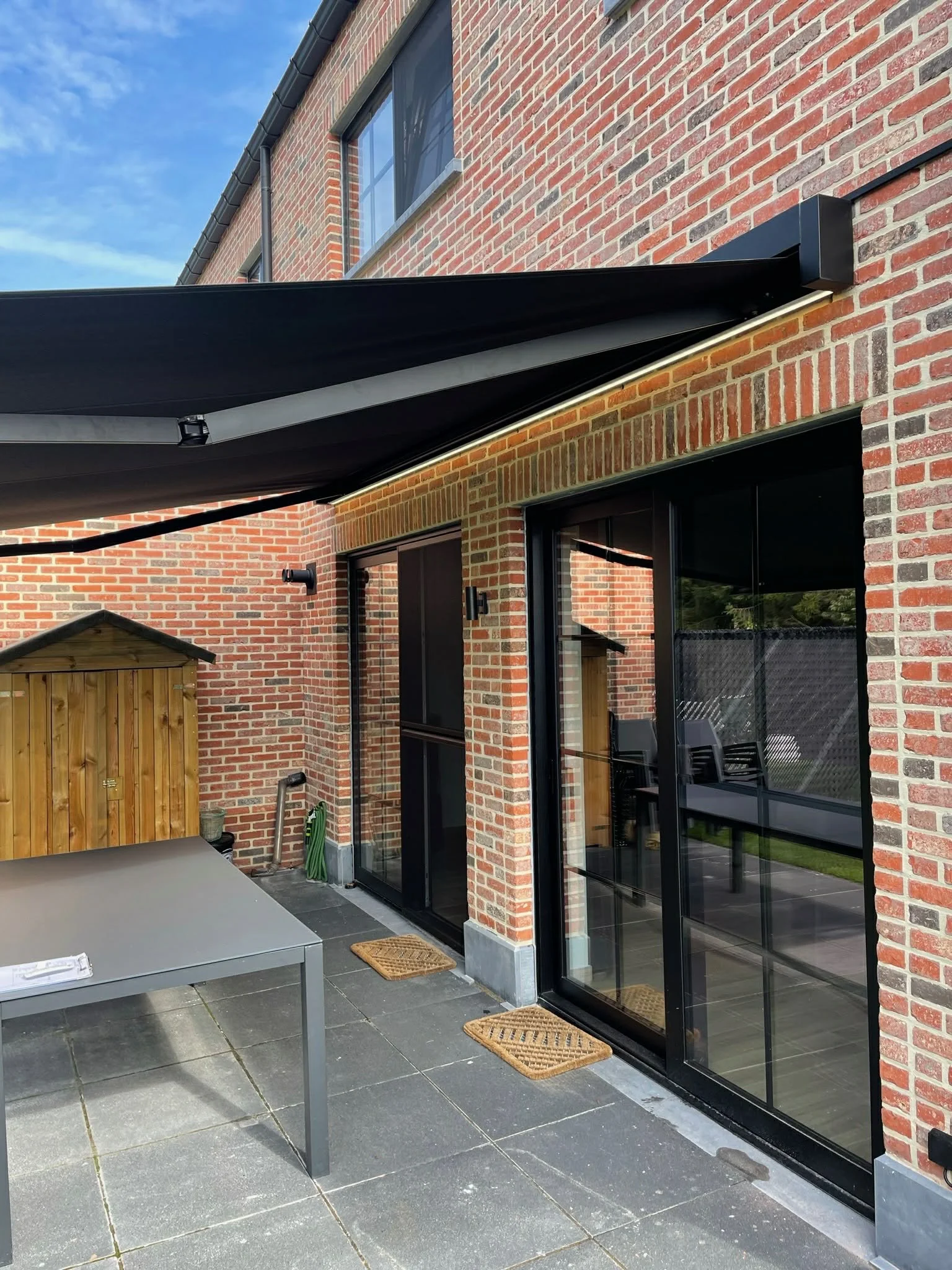 Back patio of a brick house with black sliding glass doors, outdoor furniture, and a wooden shed, with a black retractable awning and potted plants.