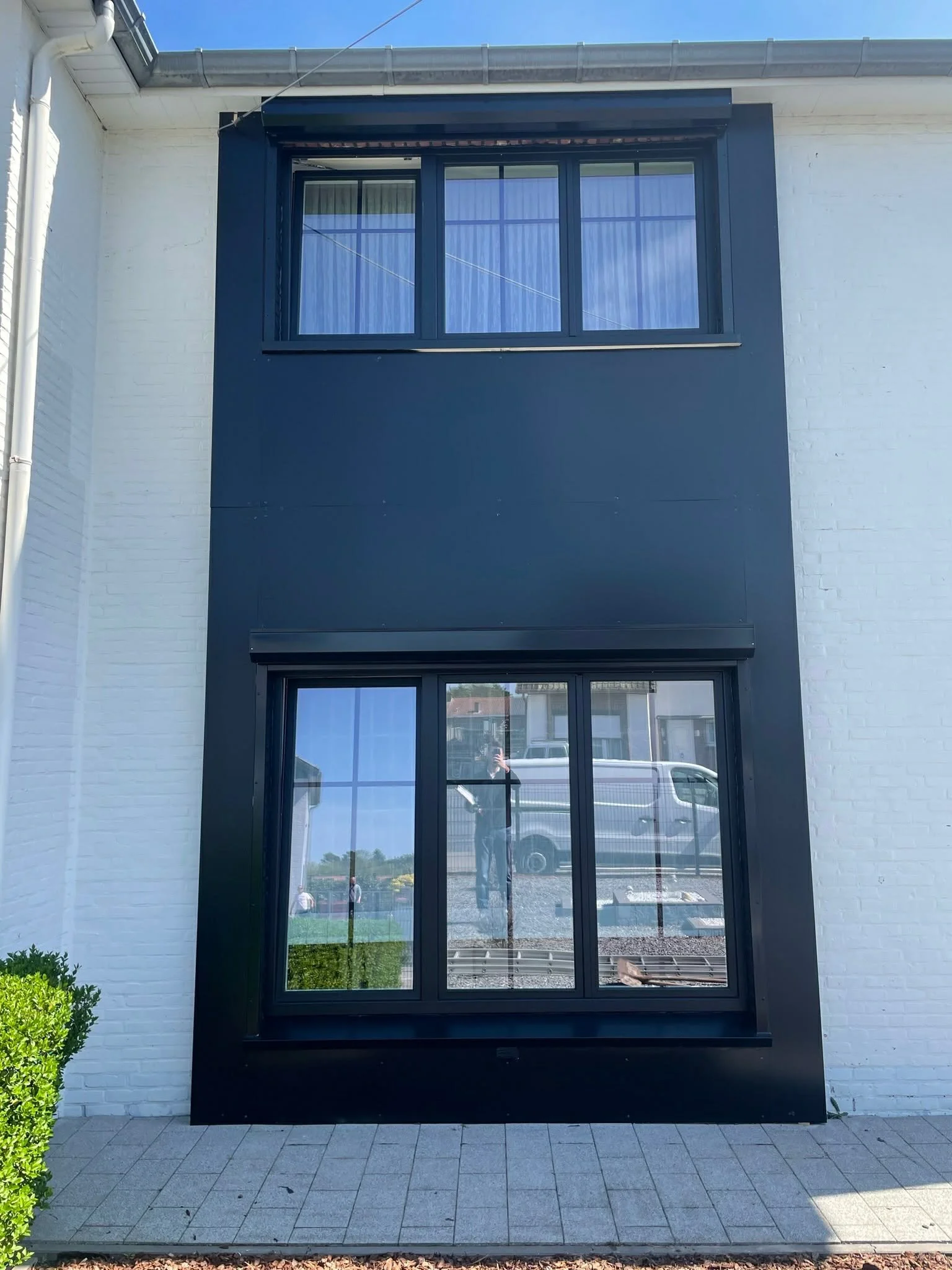 A modern building exterior with two large rectangular black-framed glass windows, one on the second floor and one on the ground floor, set against a white brick wall. The windows reflect the outdoors, including a person taking the photo, a vehicle, a