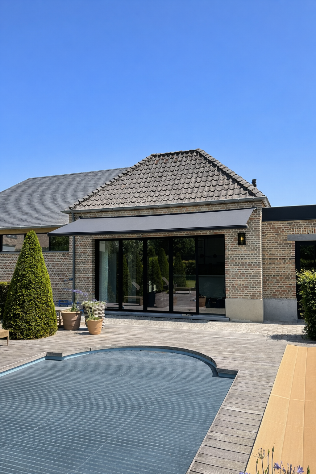 A modern backyard with a brick house, large glass doors, a pool with a cover, and a wooden deck with potted plants and outdoor furniture.