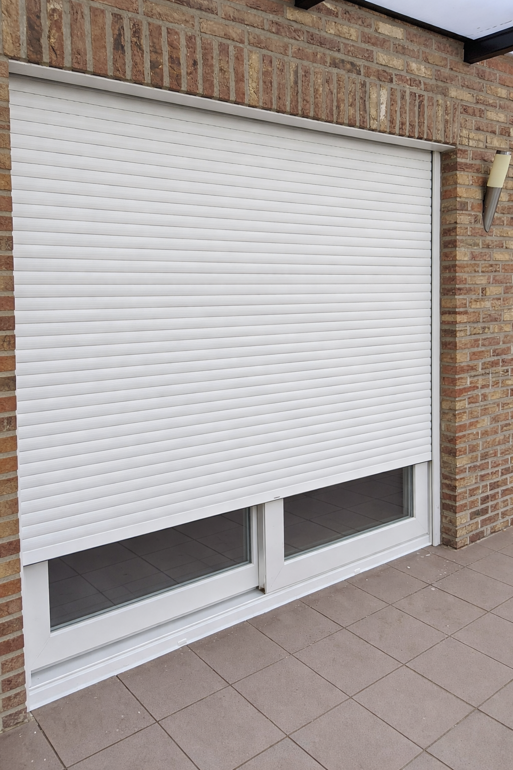 Closed white roller shutter door with glass panels at the bottom, set in a brick wall, with a wall-mounted outdoor light fixture to the right.