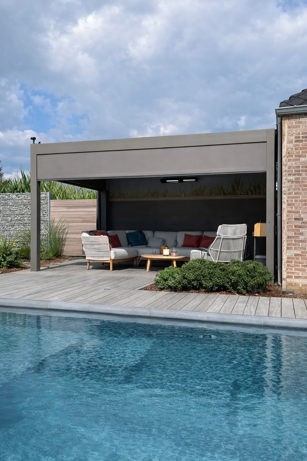 Poolside outdoor patio with modern lounge seating under a canopy, surrounded by plants and a brick wall, with a cloudy sky overhead.
