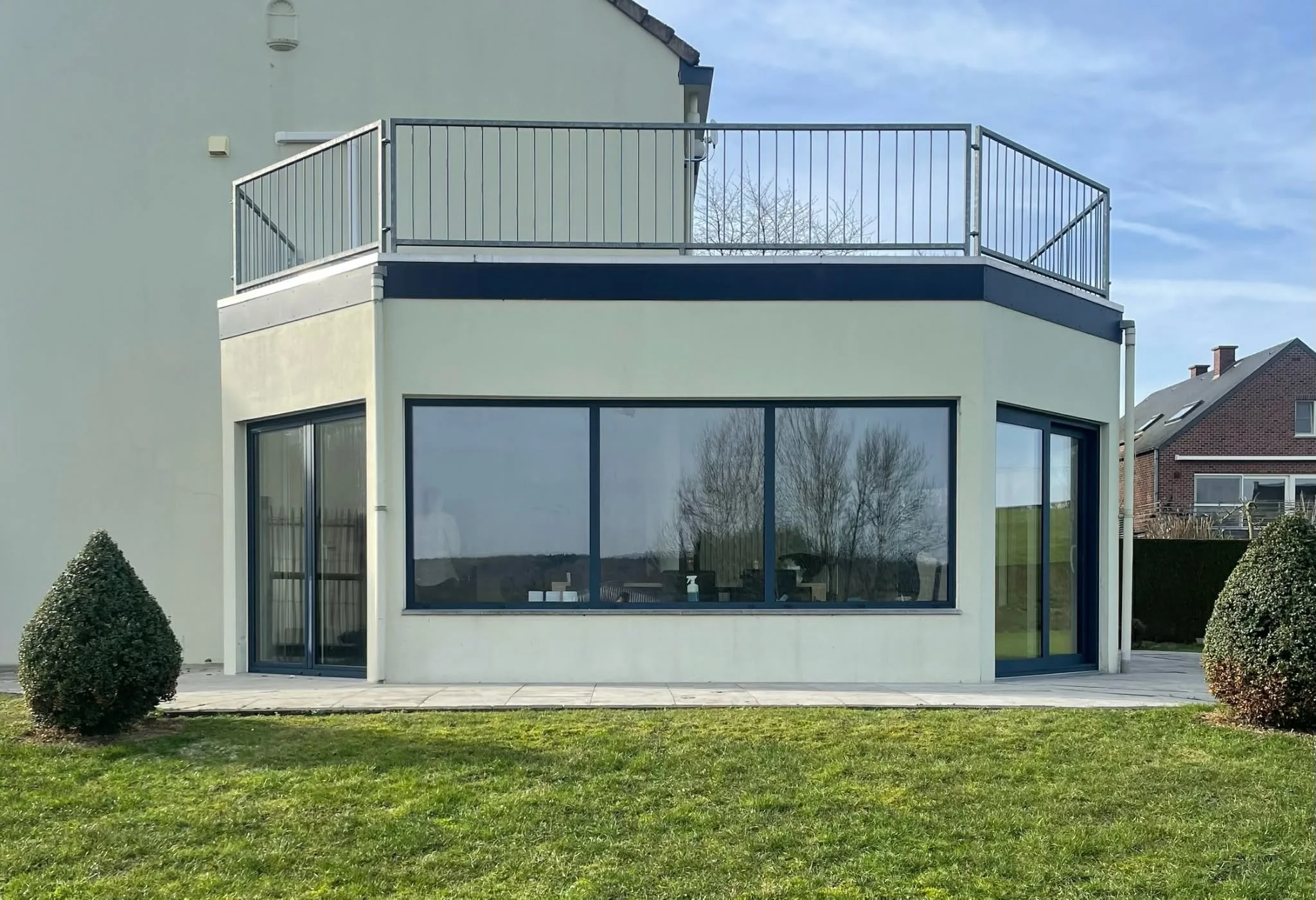 Modern house with large glass windows and sliding doors on the ground floor, a balcony with metal railing on the upper floor, and a well-maintained lawn with two bushes in front.