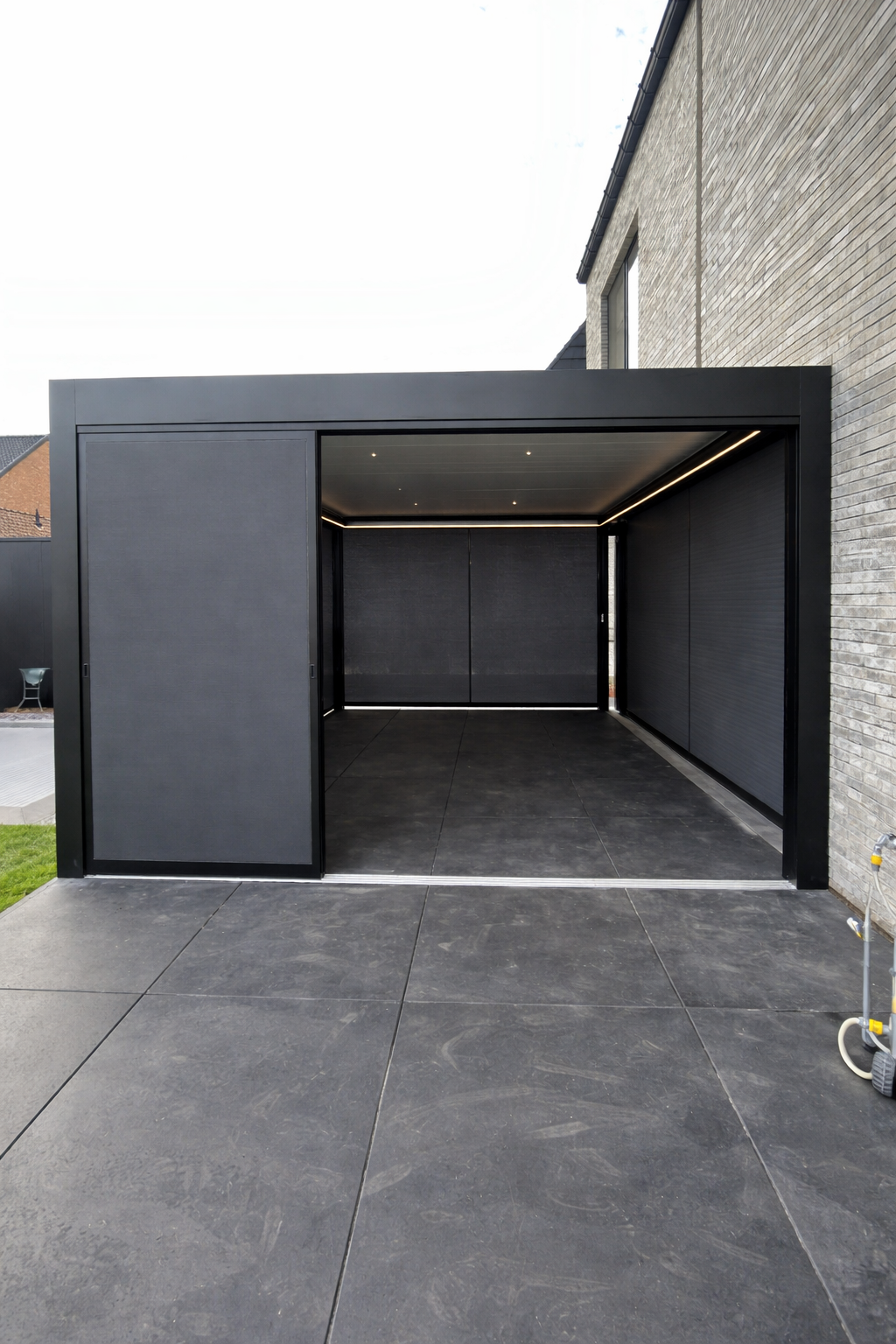 Modern black garage with sliding doors, open to a paved patio area, attached to a brick house.