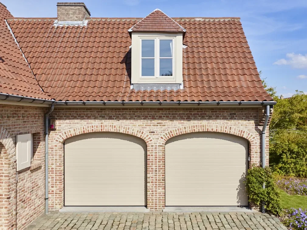 Two beige garage doors on a brick house with a red-tiled roof and a dormer window above.