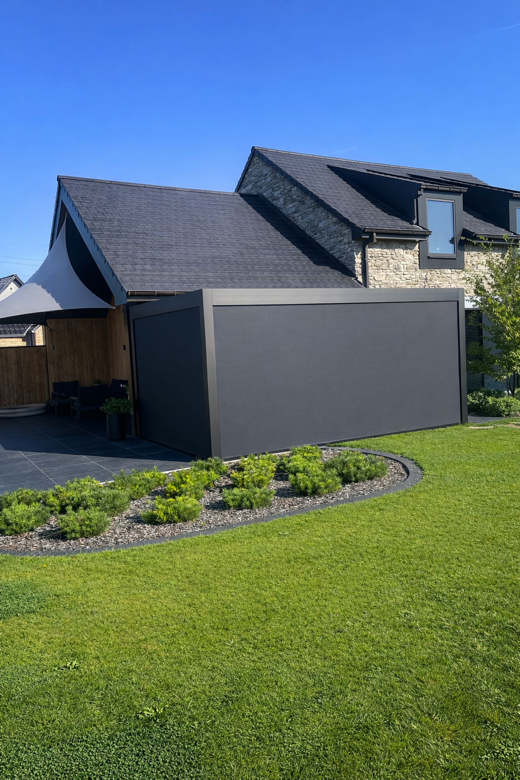 Modern house with a gray tiled roof, stone and siding exterior, and a shaded patio area with outdoor seating. Front yard has a well-maintained lawn and a flower bed with small green plants.