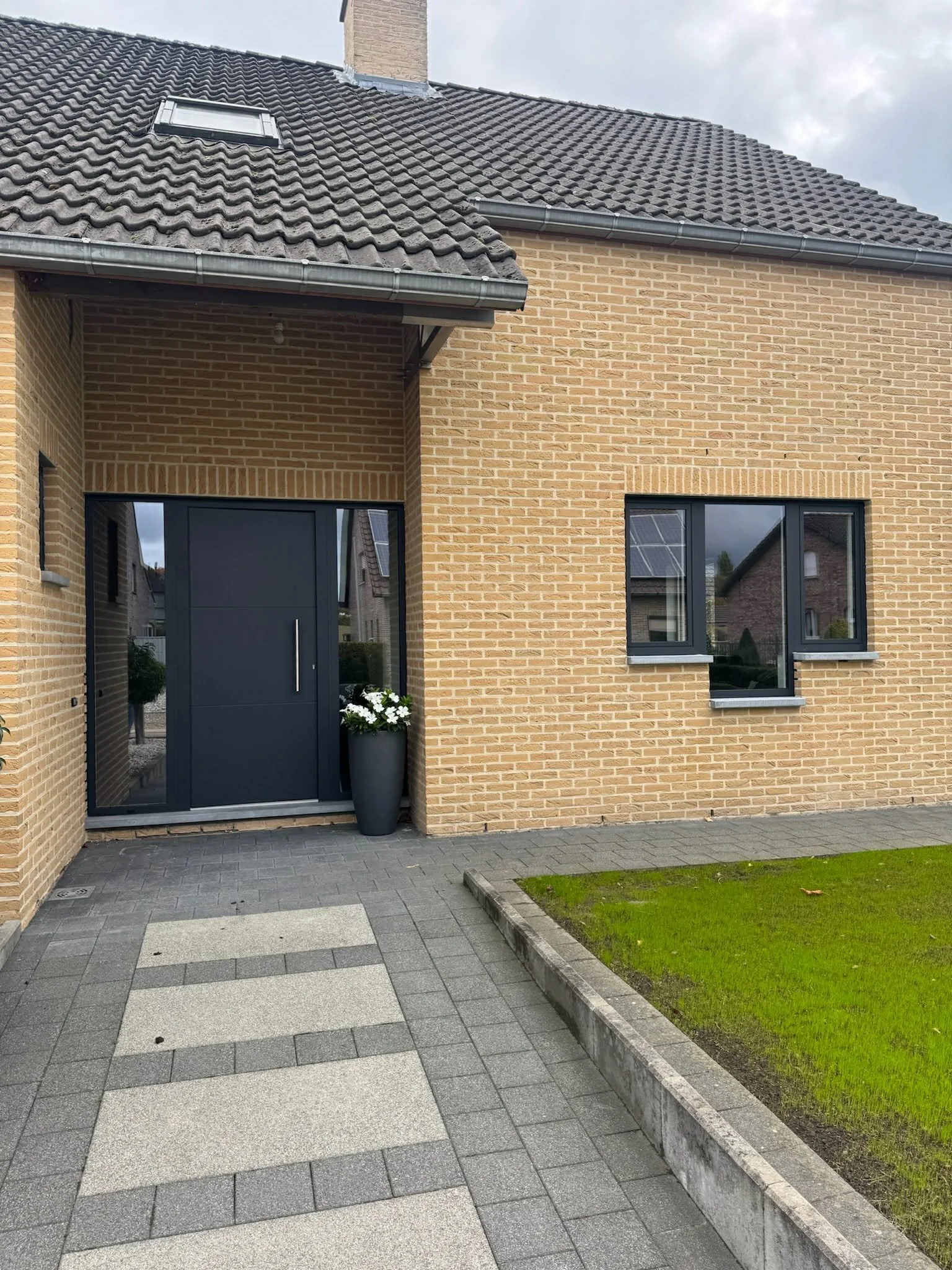 Modern house entrance with dark gray door, large gray planter with white flowers, brick exterior walls, and gray tiled pathway leading to the front door, with lawn on one side and windows reflecting neighboring houses.