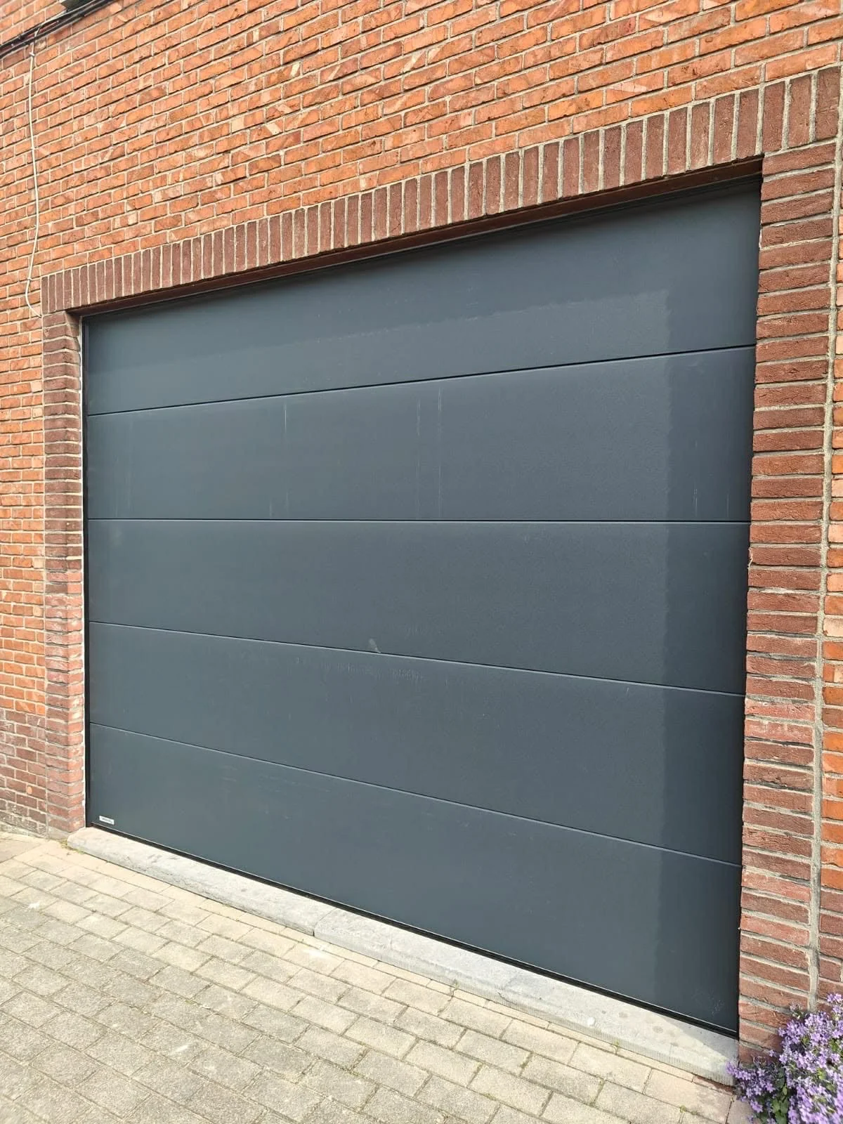 A closed gray garage door set within a red brick wall. The ground in front has gray paving stones and there are purple flowers in the bottom right corner.