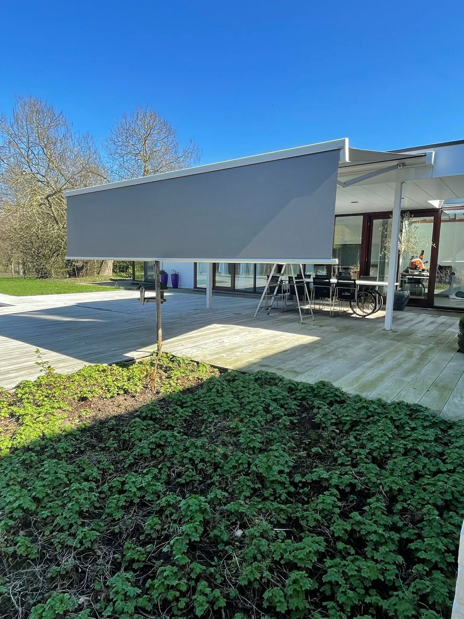 Modern house exterior with a wooden deck, large windows, and an adjustable grey awning. There is outdoor furniture, a bicycle, and potted plants on the patio. A garden with green foliage is in the foreground, and leafless trees are in the background 