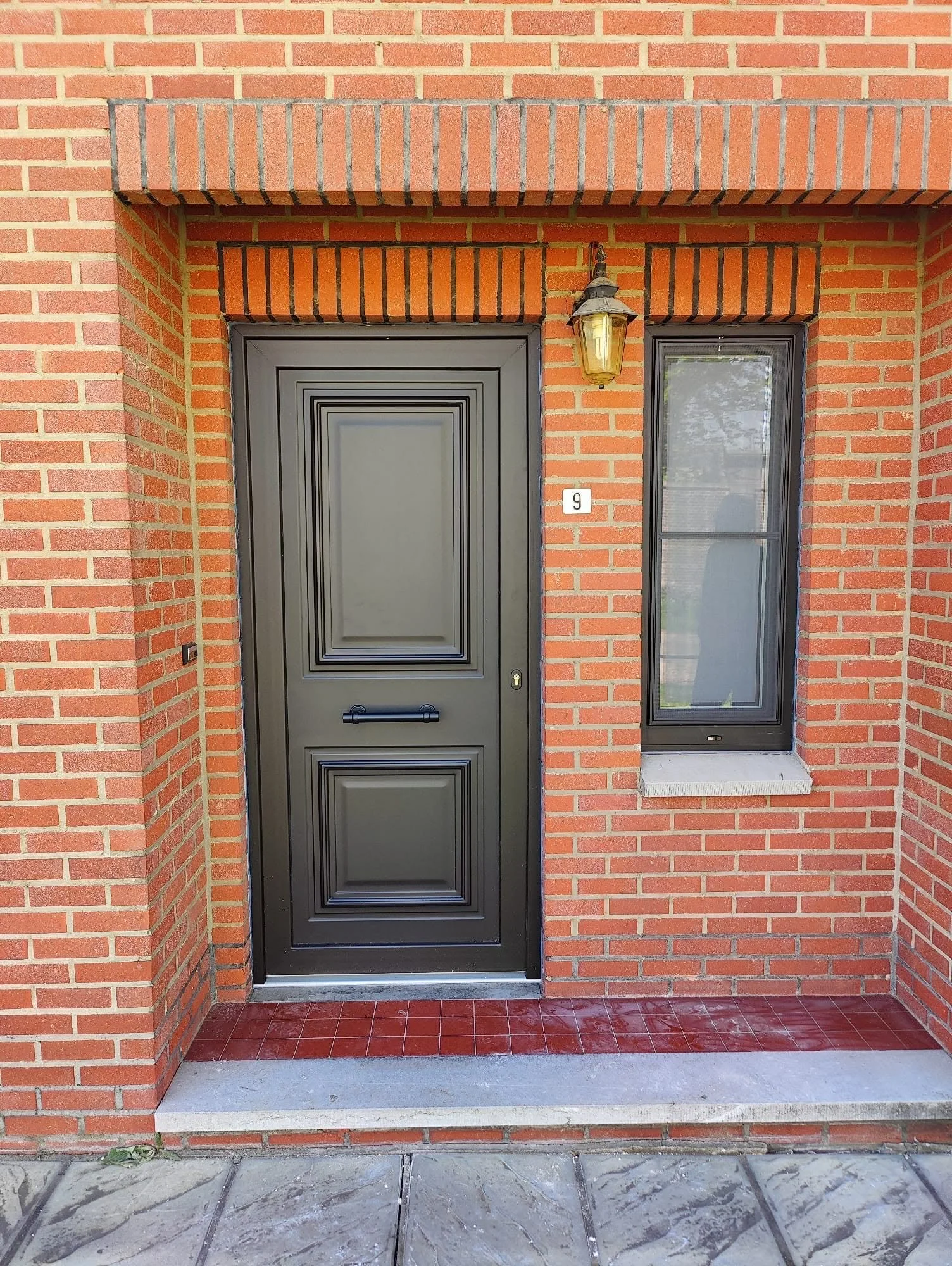 Black front door with decorative panels, to the right a tall window with black frame, brick exterior, outdoor light fixture above the door, house number 9 beside the window, brick and tile porch area.