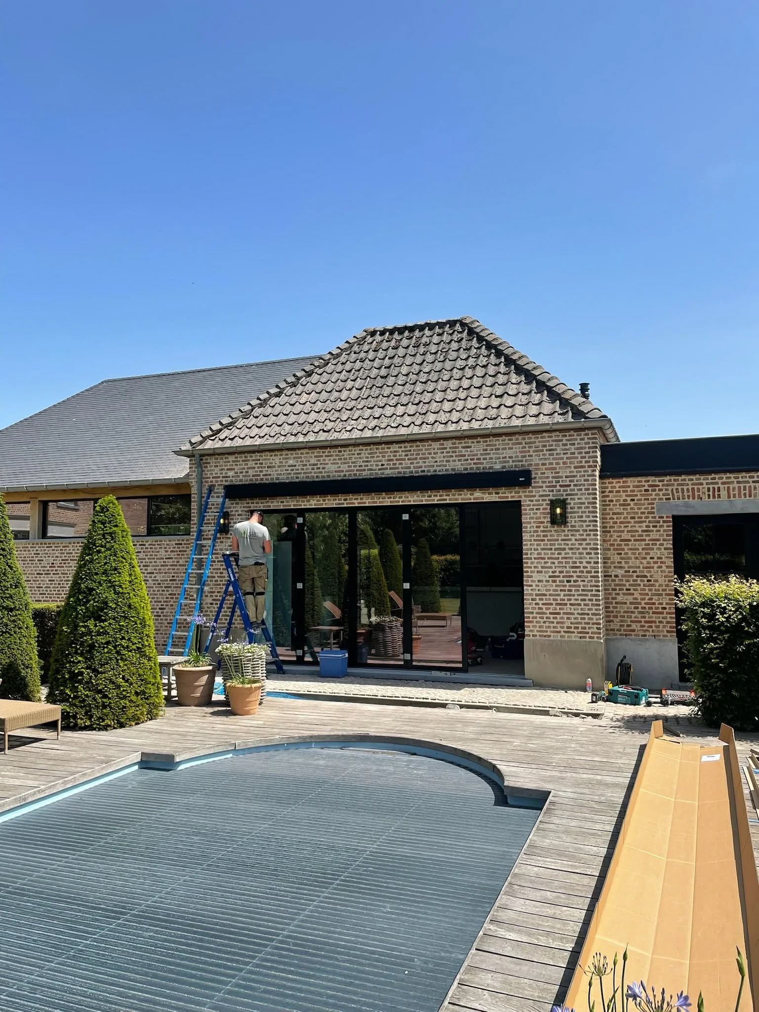 A man standing on a ladder working on the exterior of a house with brick walls and glass sliding doors, next to a backyard pool with a cover, in a residential neighborhood under a clear blue sky.