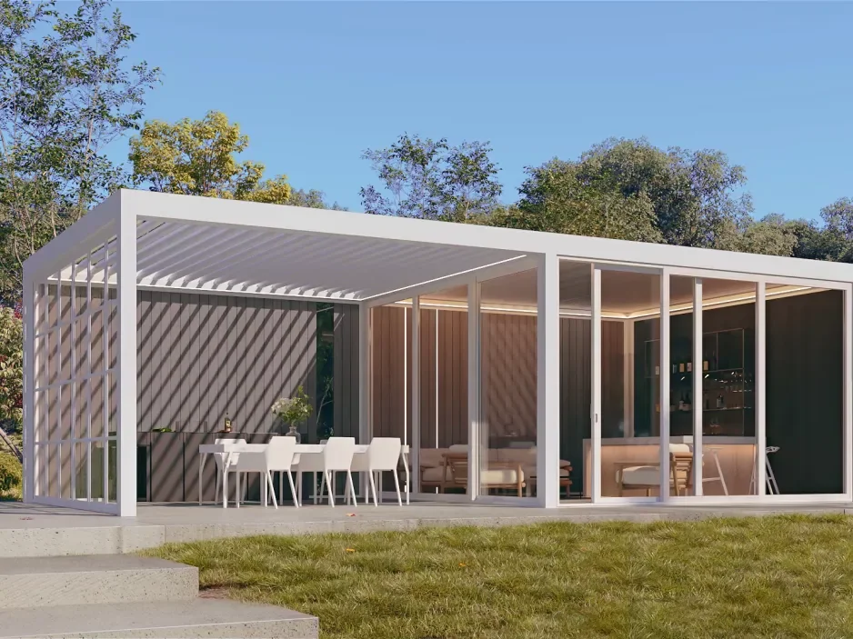 Modern glass house with white framing, outdoor dining area with white chairs, and a lounge space inside, set in a grassy yard with trees and blue sky.