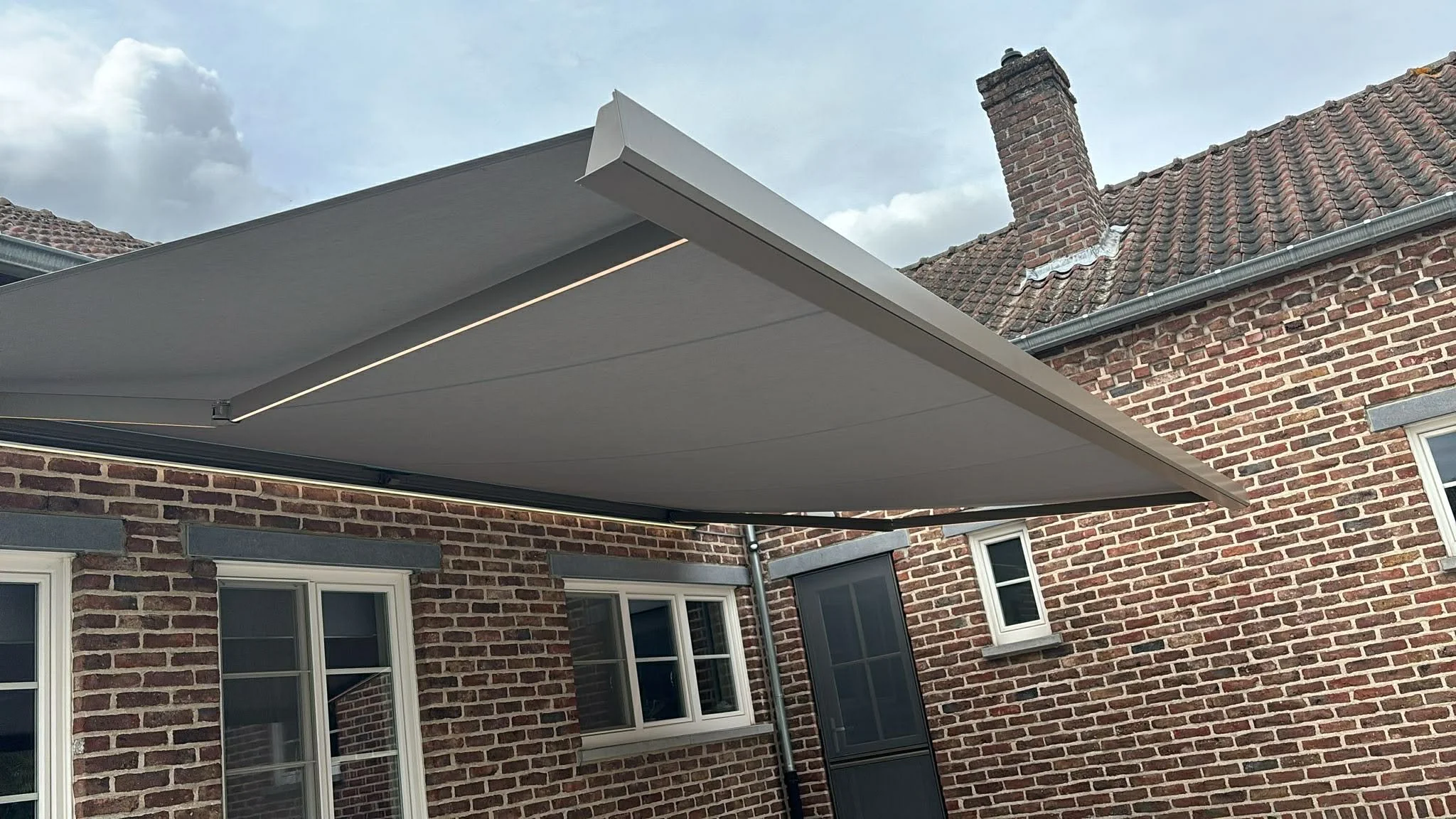 A beige retractable awning extended over a brick house patio with two windows and a screened door, with a cloudy sky above.