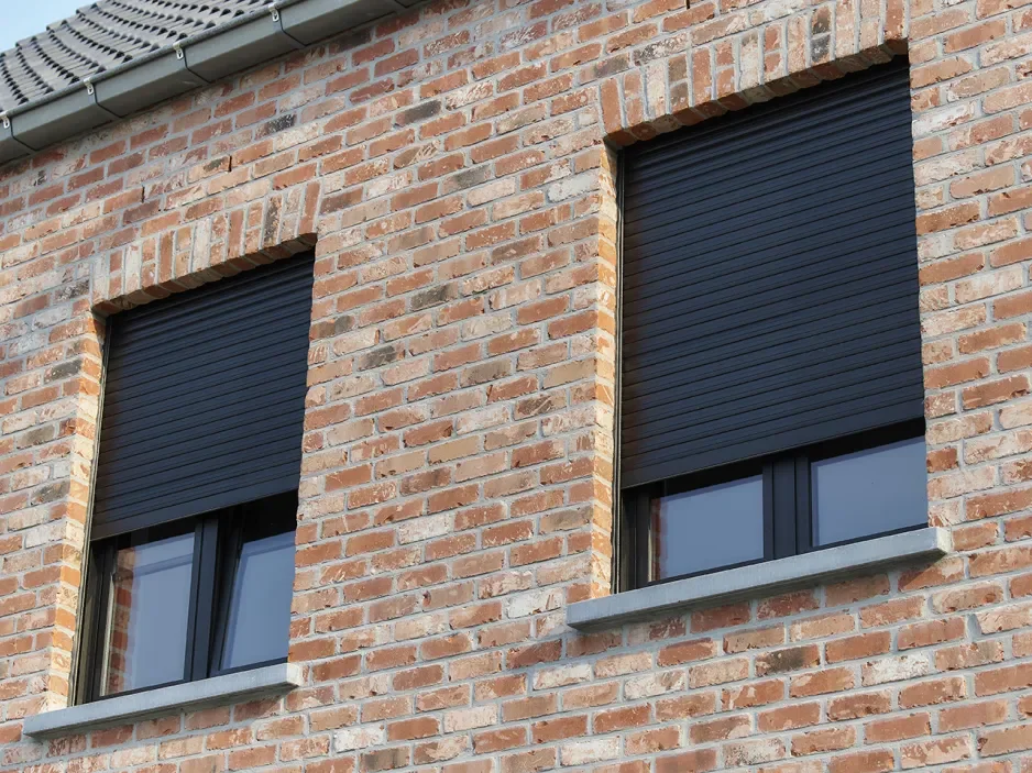 Close-up of a brick building facade with two rectangular windows featuring black rolling shutters.