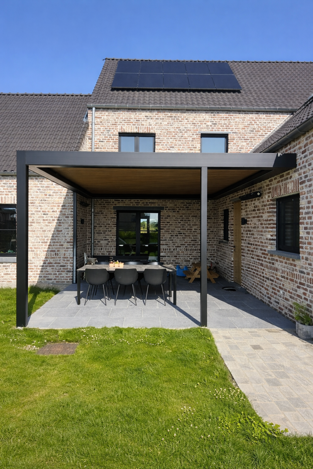 A backyard patio with a modern black pergola attached to a brick house, featuring outdoor dining table and chairs, with a green lawn and clear blue sky.