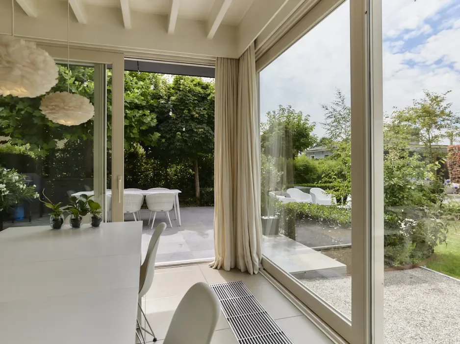 View from inside a bright, modern dining area looking out to a lush outdoor garden with trees, patio furniture, and a cloudy sky
