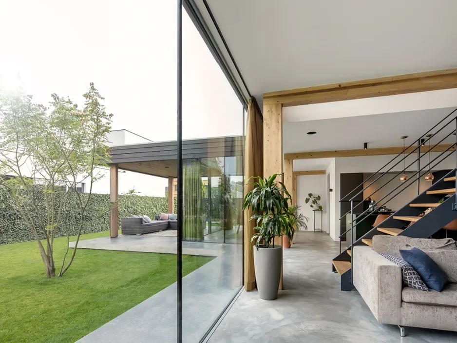 Inside view of a modern home with concrete floors, wooden beams, a staircase with black railing, a sofa with pillows, and large glass sliding doors leading to an outdoor patio with grass, a tree, and outdoor seating.