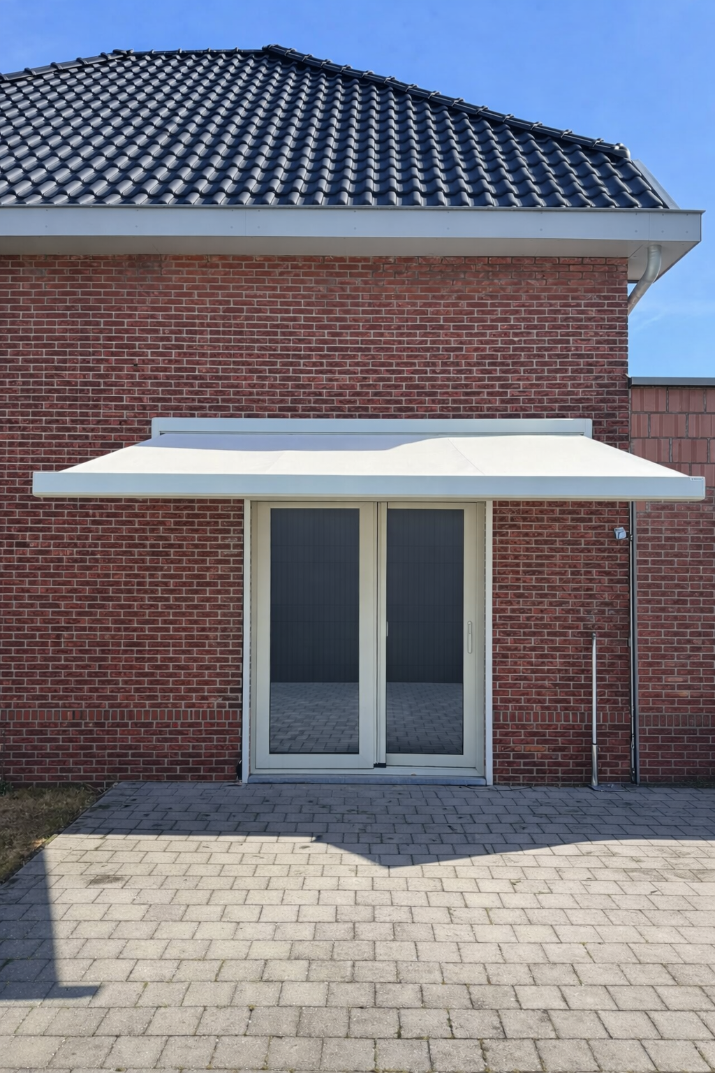 The image shows the exterior of a brick house with a tiled roof, a white sliding glass door, a white awning above the door, and a paved patio area in front.
