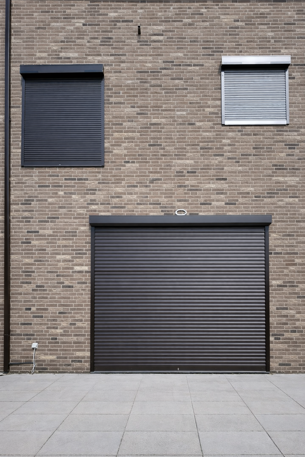 Brick building with three black metal shutters on upper and ground levels, sidewalk in front, and electrical outlet on lower wall.