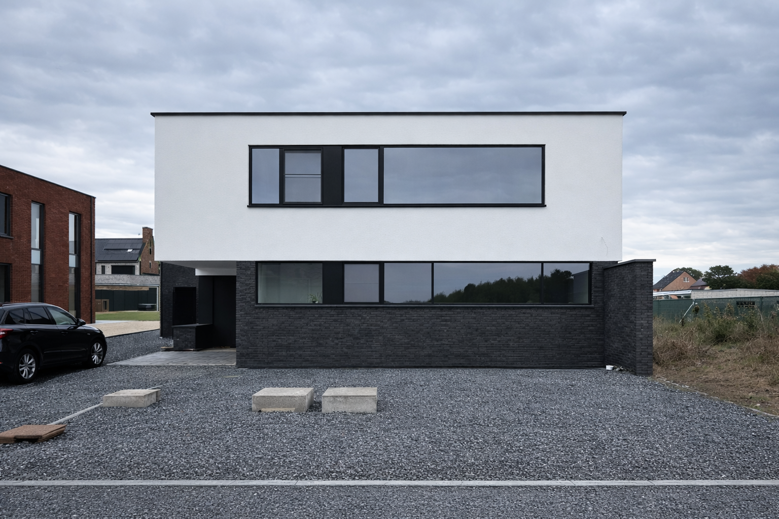 Modern two-story house with white upper and black brick lower sections, large horizontal windows, and a gravel parking area with a black car on the left.