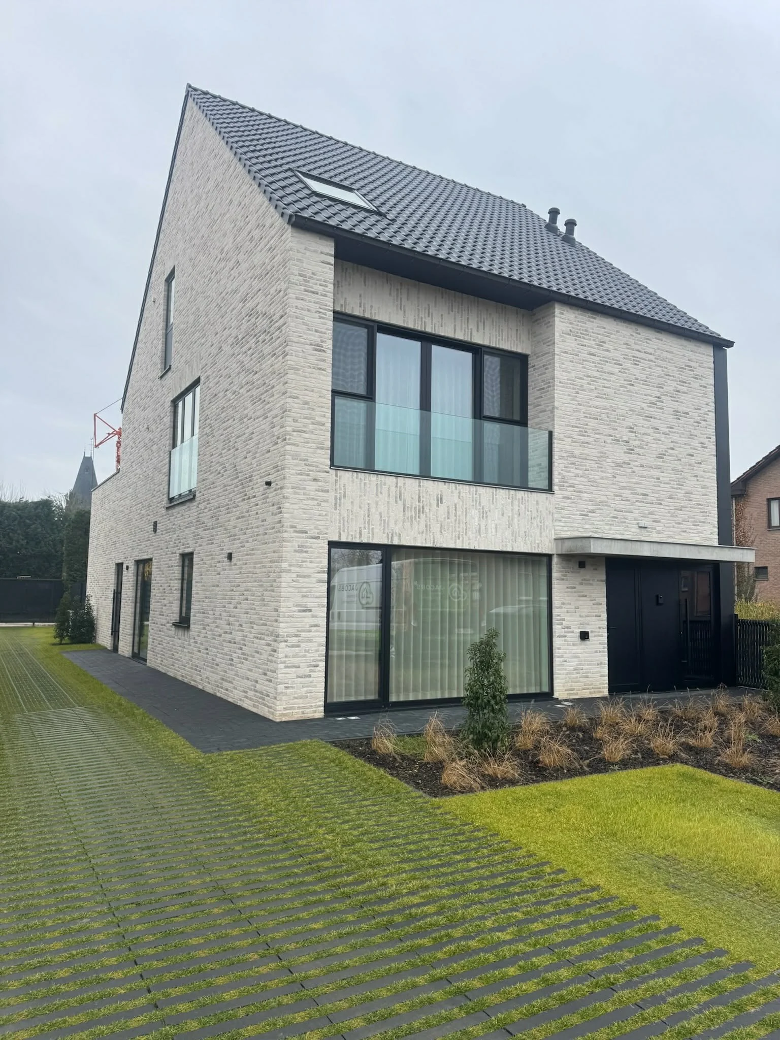 Modern multi-story house with brick exterior, large windows, and a dark roof, surrounded by a landscaped yard with grass, small plants, and paved walkways.
