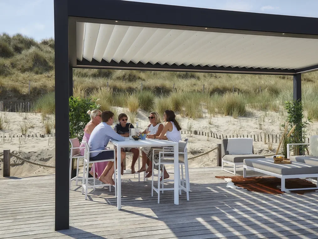 Group of five people enjoying a meal at an outdoor table on a beach deck with a sandy beach and grassy dunes in the background.