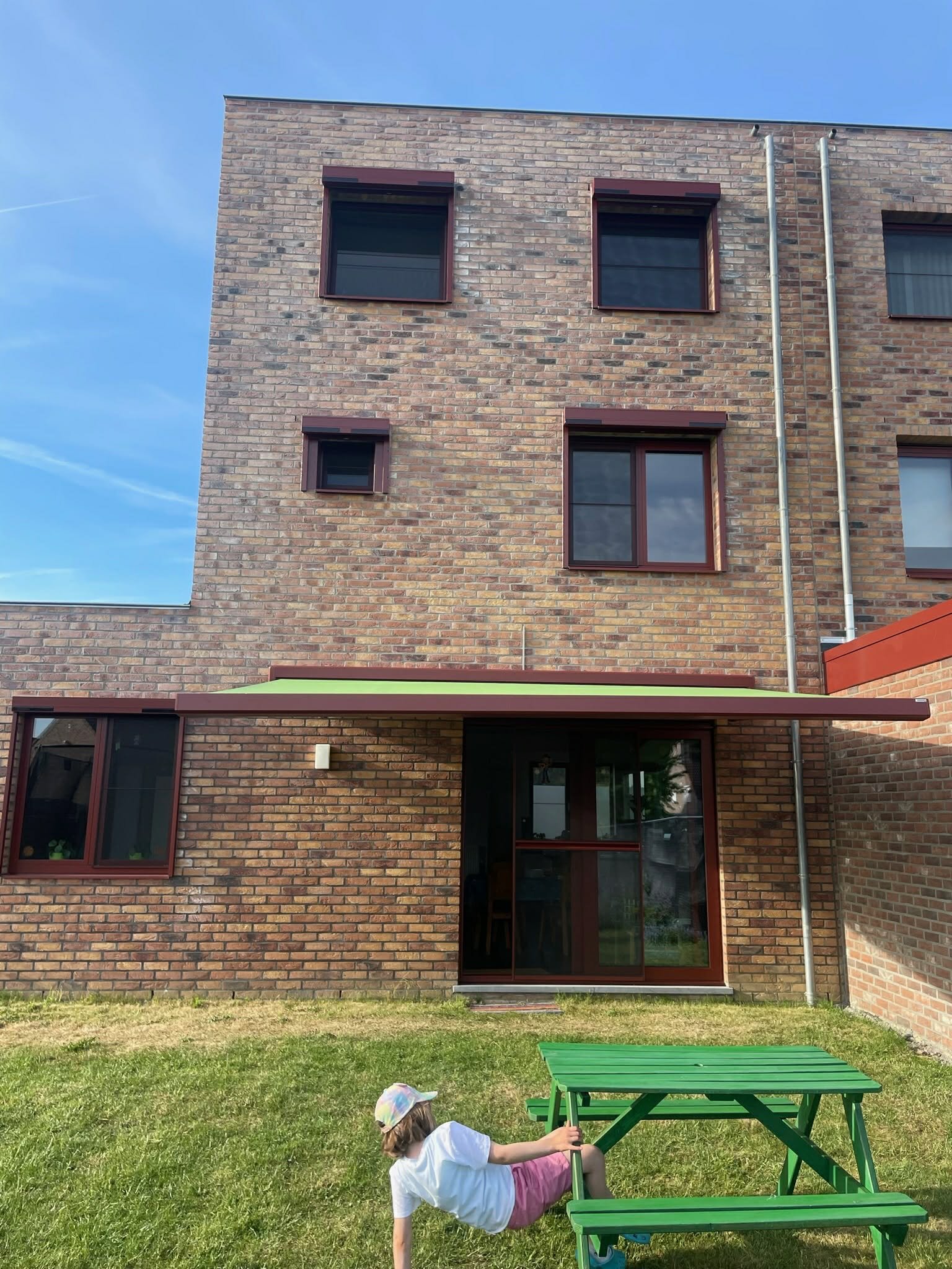 A child lying on the grass near a green picnic table in front of a brick building with multiple windows and a sliding glass door.