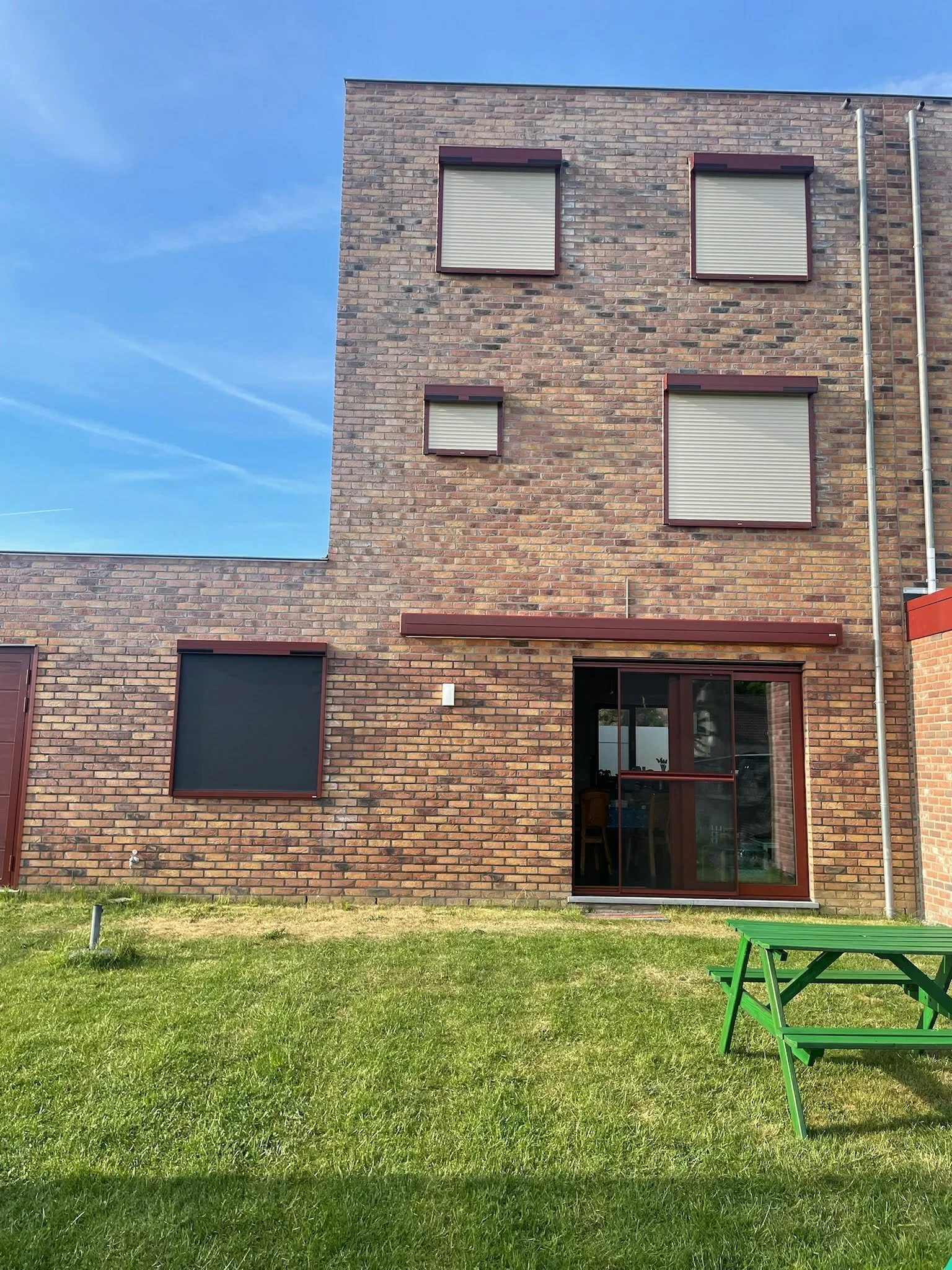 A multi-story brick residential building with several windows, some with closed blinds, a sliding glass door, a grassy yard, and a green picnic table outside under a blue sky.