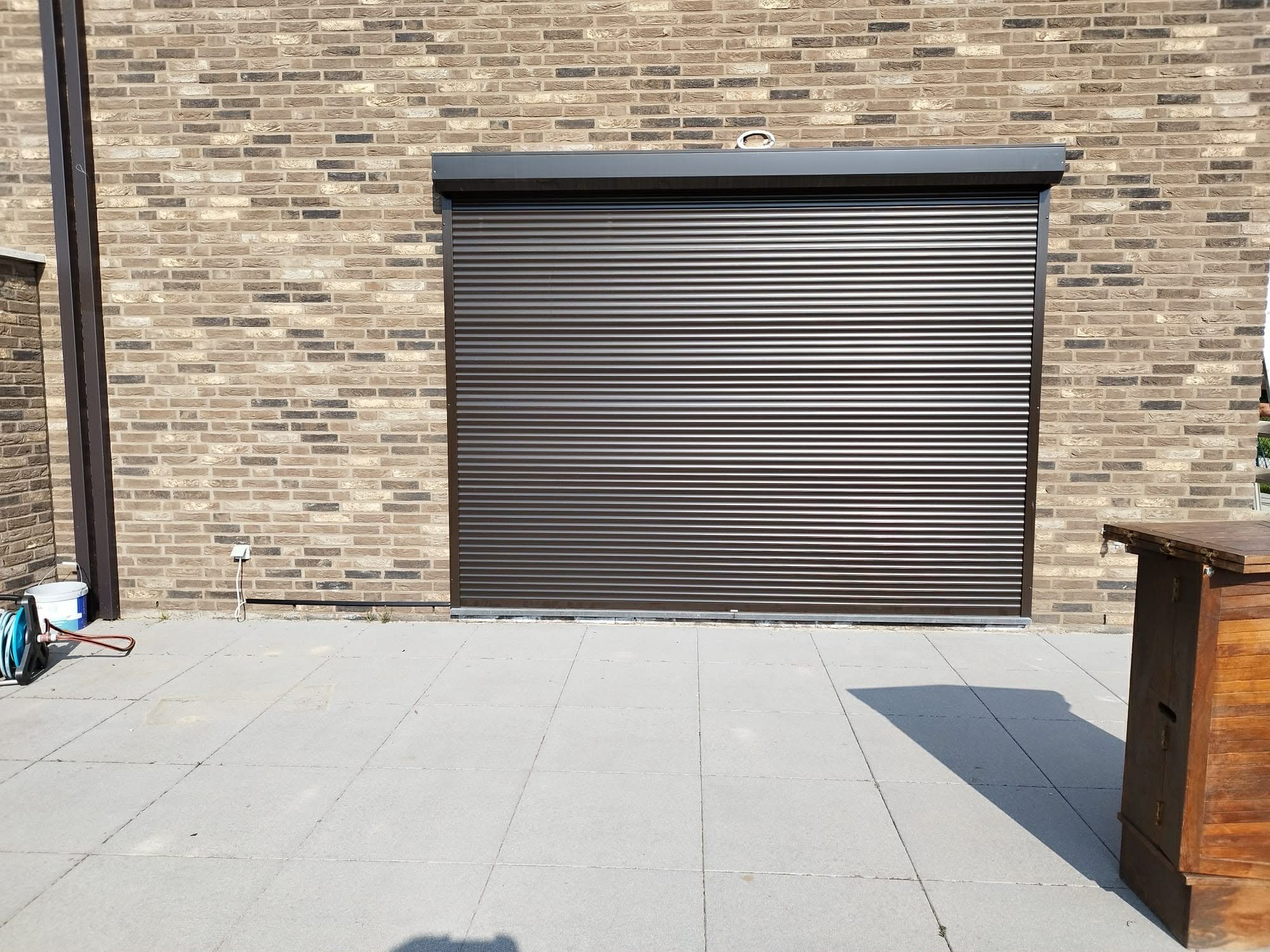 Steel rolling door on a brick wall exterior, with a concrete patio in front and objects on the left and right sides.