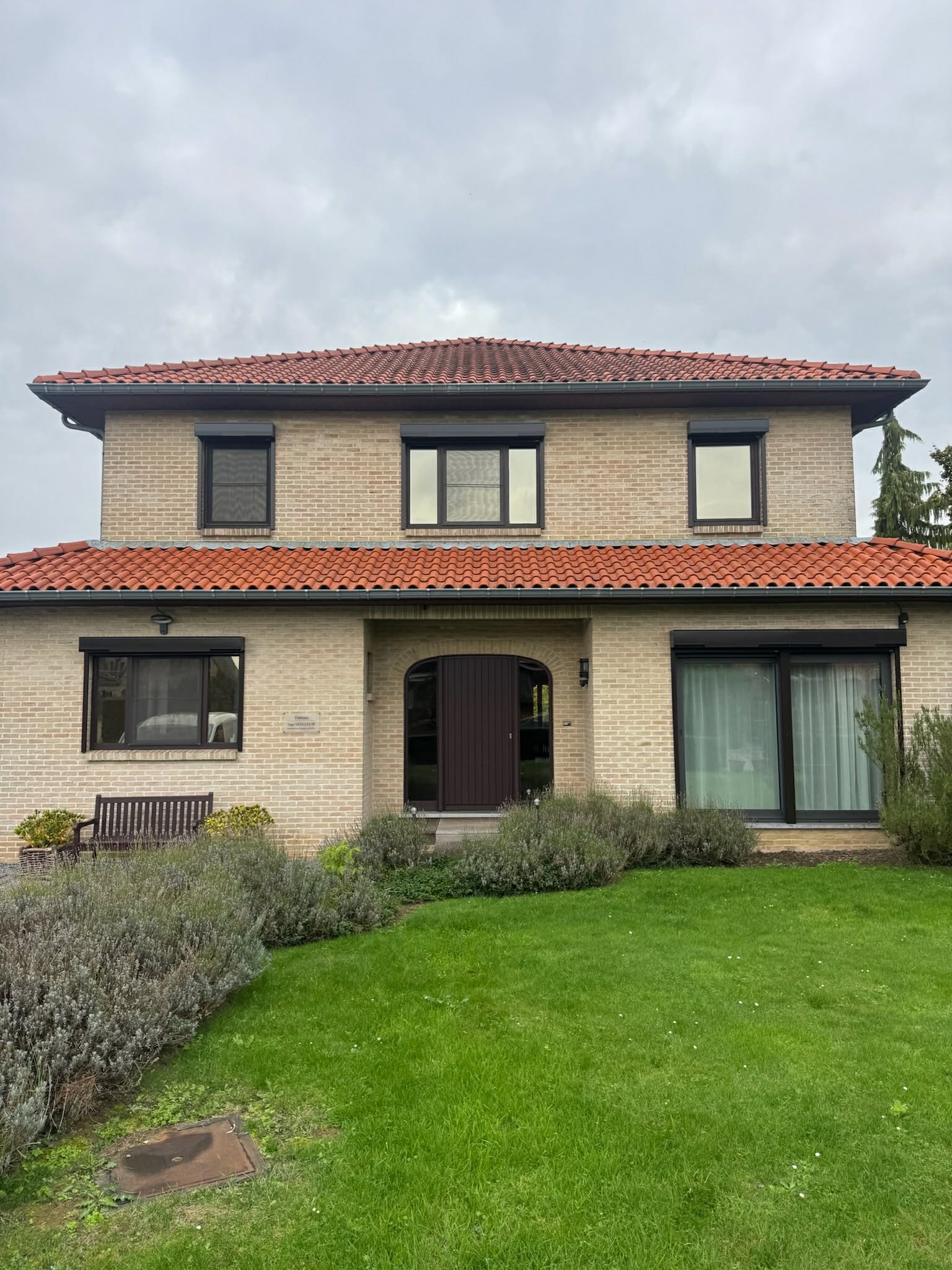 A two-story house with beige brick walls, black window frames, and a red tiled roof. There is a garden with green grass, bushes, and a wooden bench in front of the house.