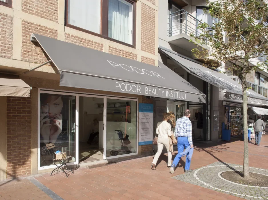 Exterior of a beauty institute shop named Podor with glass doors and a gray awning, in a shopping area with people walking by and a small tree in front.