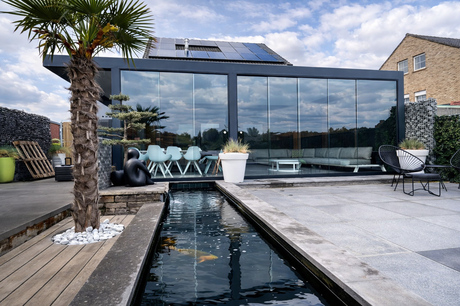 Modern rooftop patio with a rectangular water feature, a large glass enclosed lounge area, and outdoor furniture including a long sofa, chairs, and tables. Potted plants and a palm tree are present, with a cloudy sky reflected in the glass.