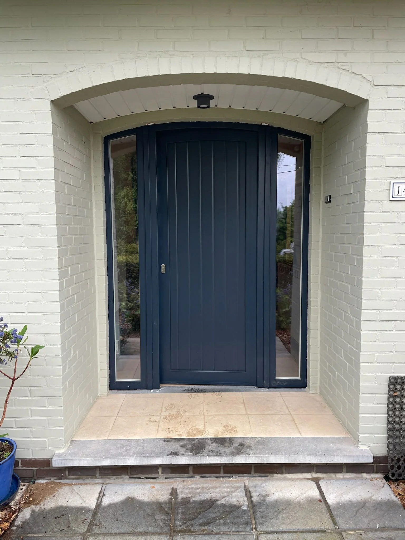 The front entrance of a house with a navy blue curved front door, flanked by glass panels, and a light-colored brick exterior. There is a small paved area and a potted plant to the left.