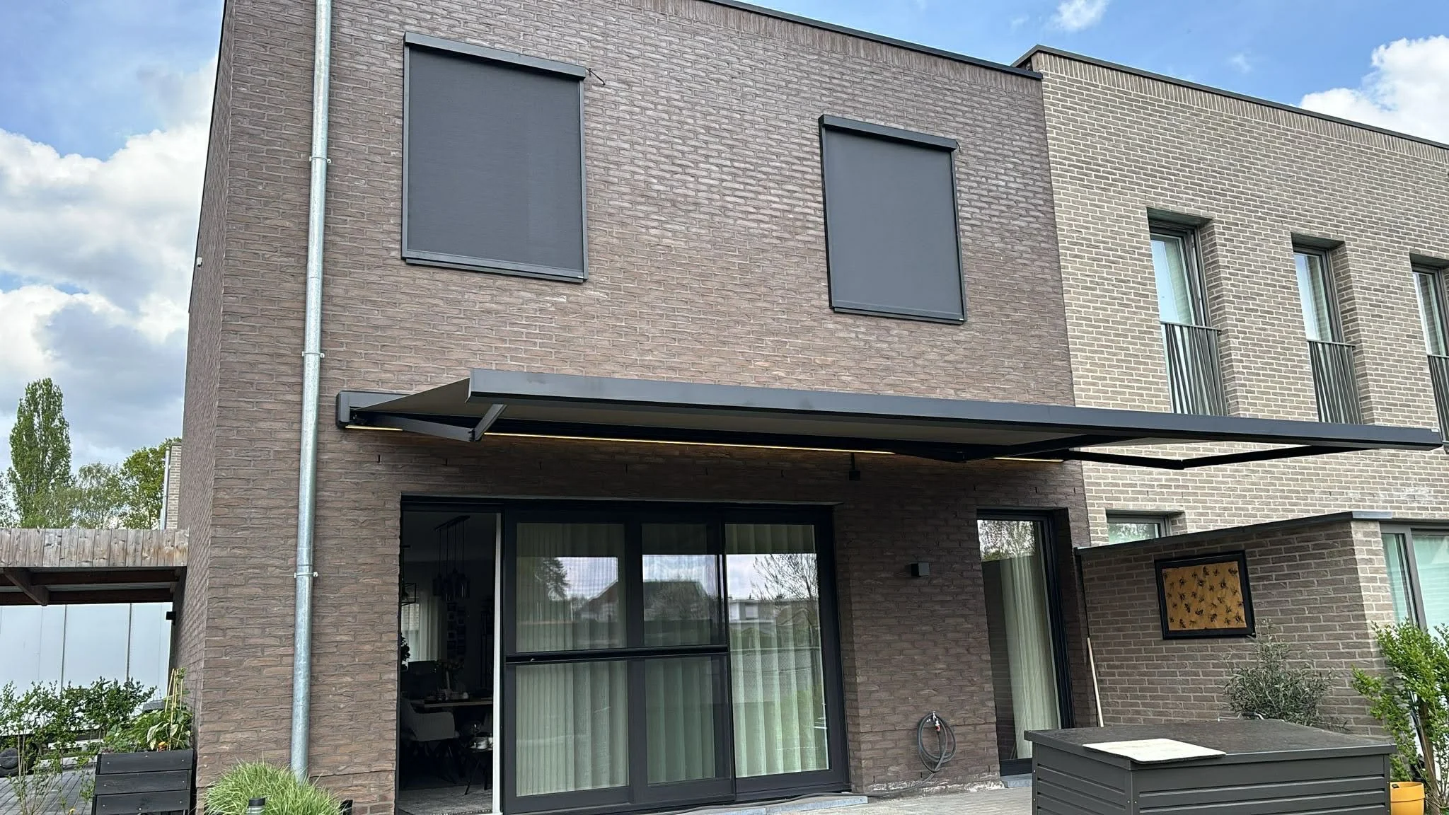 Modern three-story residential building with brick exterior, large glass sliding door, and window shutters closed on two windows.