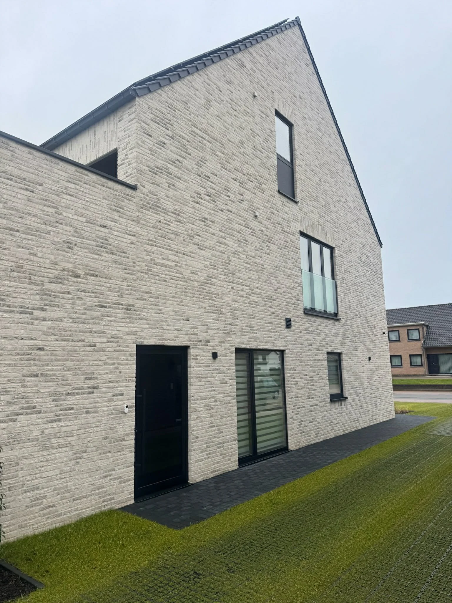 Modern multi-story house with a light gray brick exterior, black doors, and large windows, surrounded by a lawn and paved pathway.