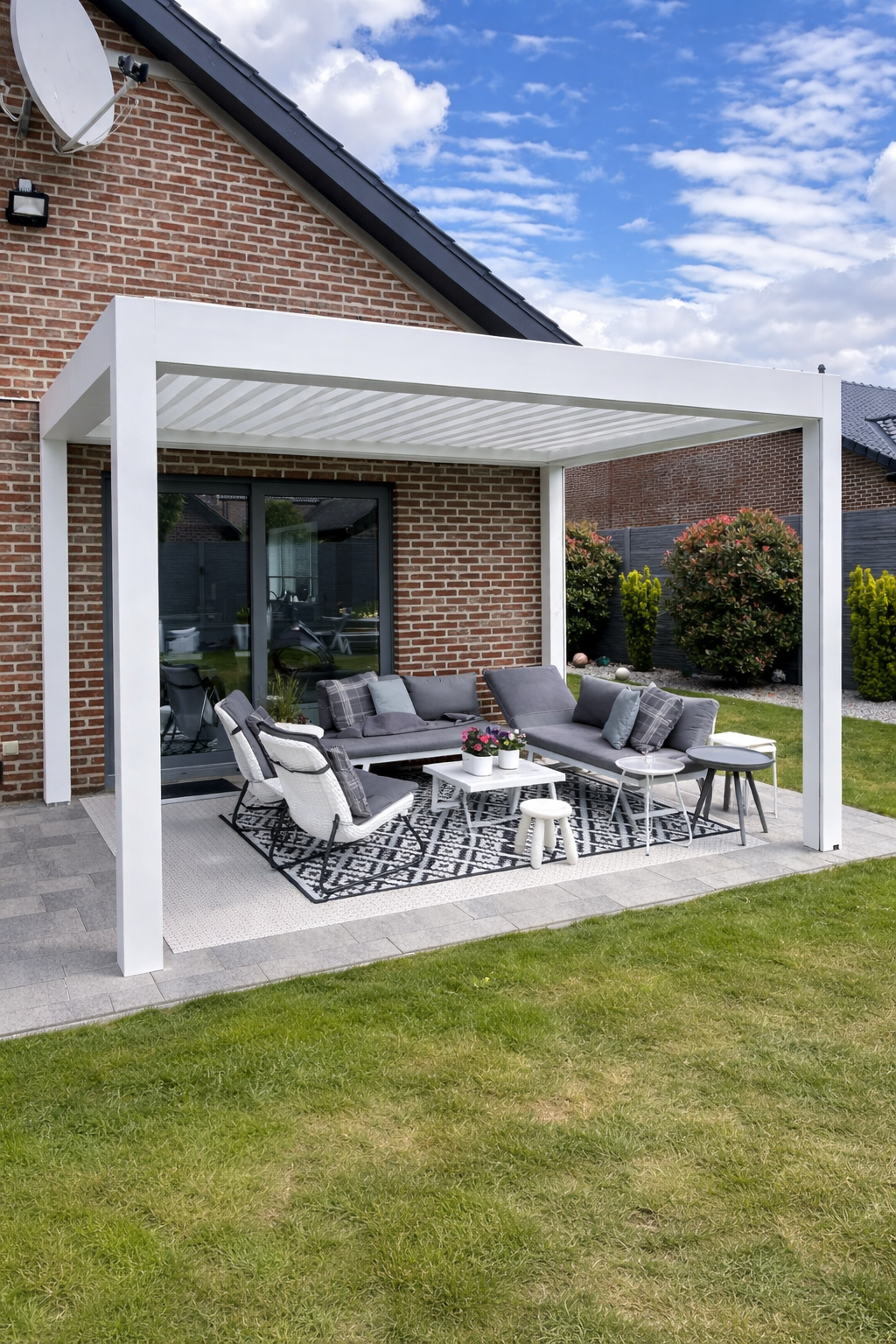 Outdoor patio with a white pergola, modern gray sectional sofa with cushions, a patterned black-and-white rug, small white and gray tables, and a grassy backyard with bushes and trees under a partly cloudy sky.