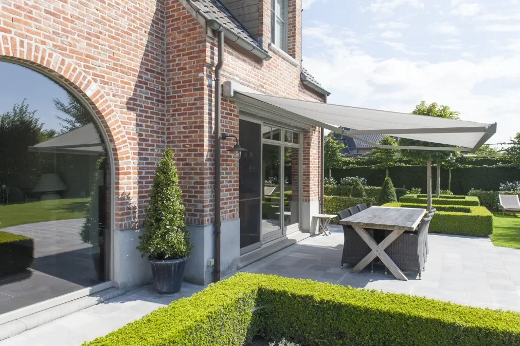 Backyard patio with a table, chairs, and an extendable shade sail, surrounded by neatly trimmed hedge in front of a brick house with large arched and rectangular windows.